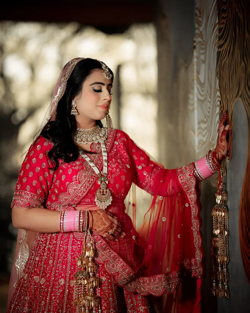 A bride in a red lehenga with kaleere rests her hand against a carved wooden screen