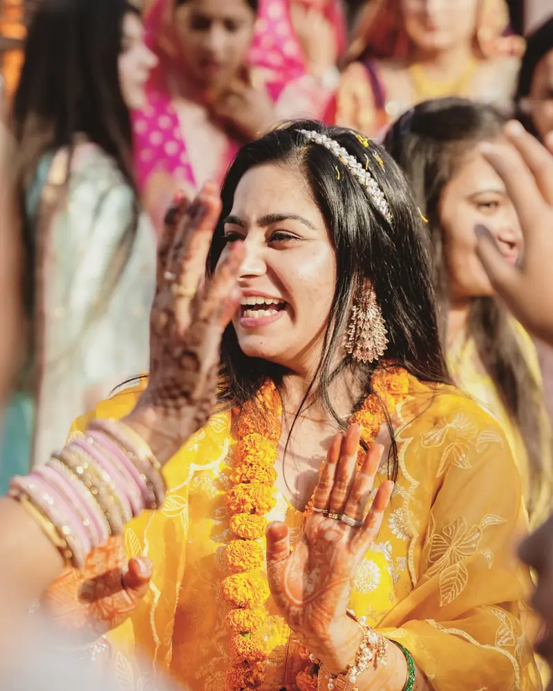 A laughing bride in yellow with a marigold garland, mehendi on her raised hands