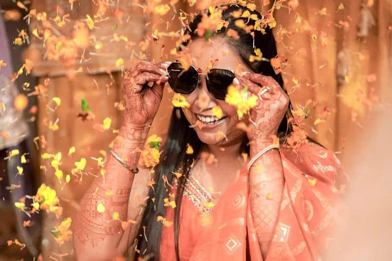 A woman in sunglasses laughs as marigold petals fall around her at a haldi