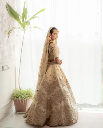 Mukta glances back over her shoulder by a tall window and a potted palm