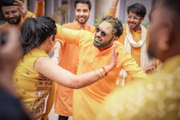A friend in yellow pushes back the groom, both laughing, turmeric on his face