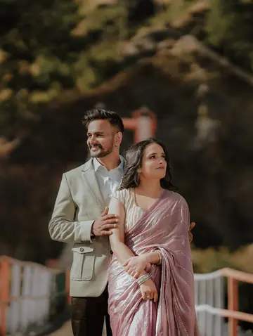 He holds her from behind, both smiling, pine trees soft behind them