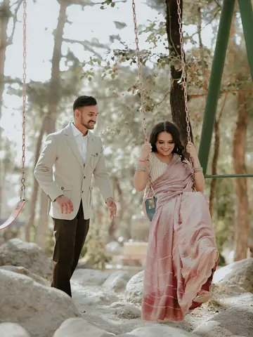 She sits on a wooden swing in a pink saree as he walks toward her through the pines