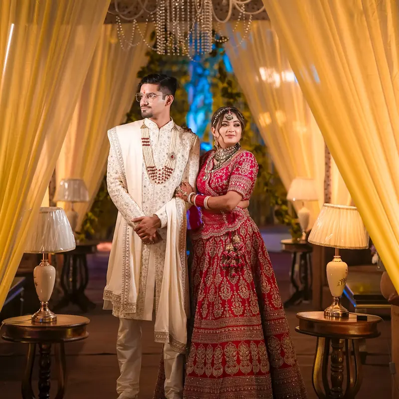Vaibhav in an ivory sherwani and Bhavya in a red lehenga stand under a gold-lit canopy of draped curtains