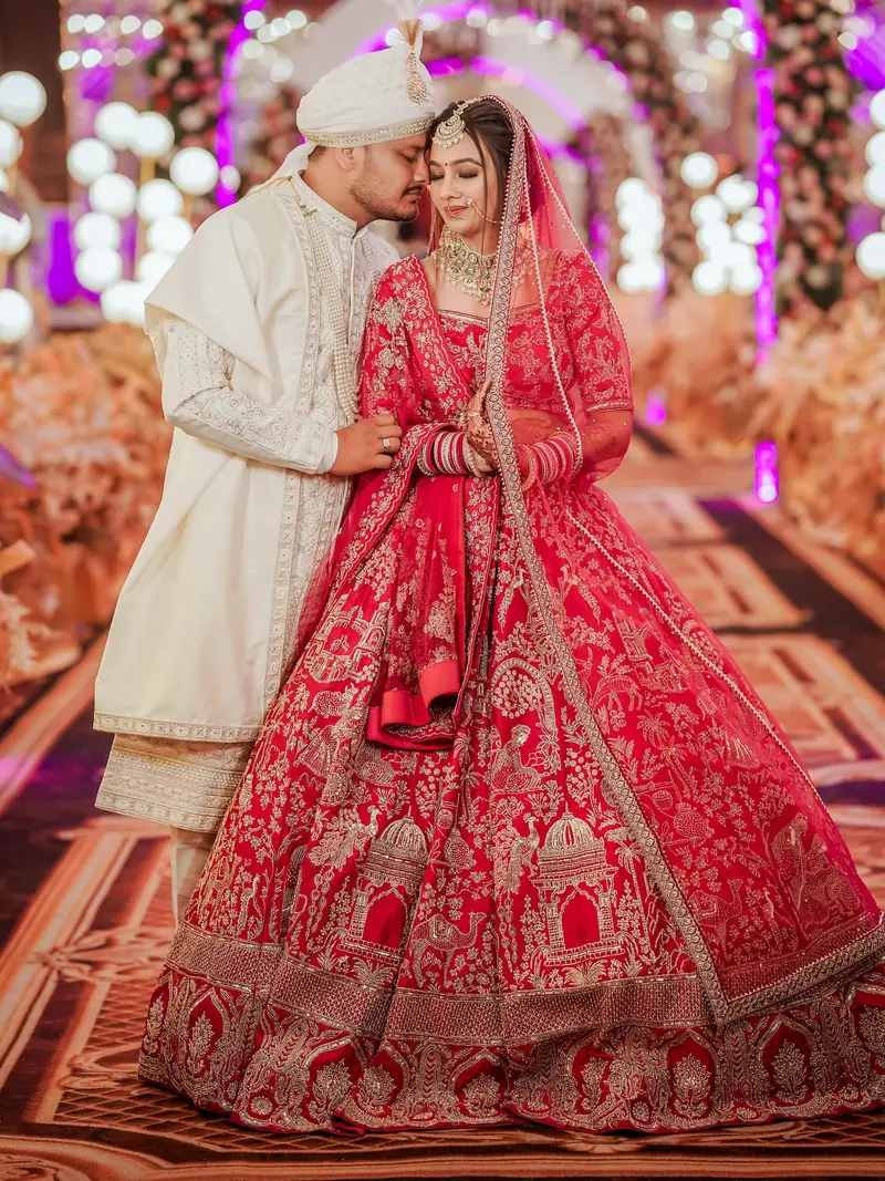 Groom in an ivory sherwani holds his bride in a red lehenga close, pink lights behind them